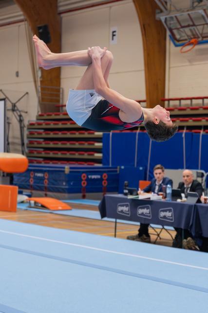 Young male gymnast performs a tucked backflip mid-air, smiling, above a blue mat at an indoor gymnastics competition.