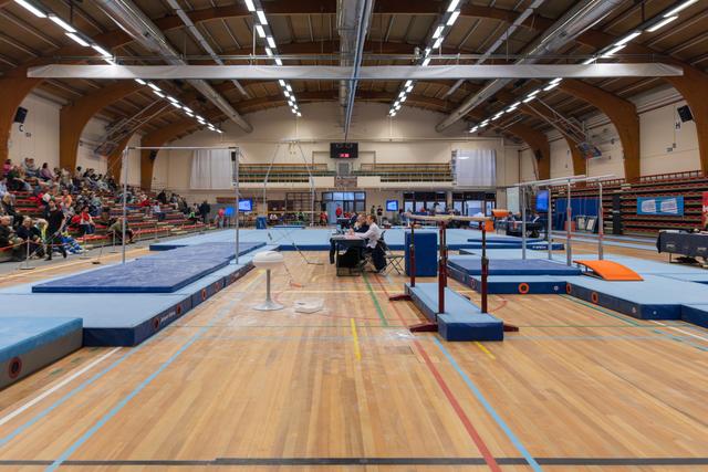 Indoor gymnastics venue with uneven bars set up, a judge seated at the scoring table, spectators filling the bleachers.