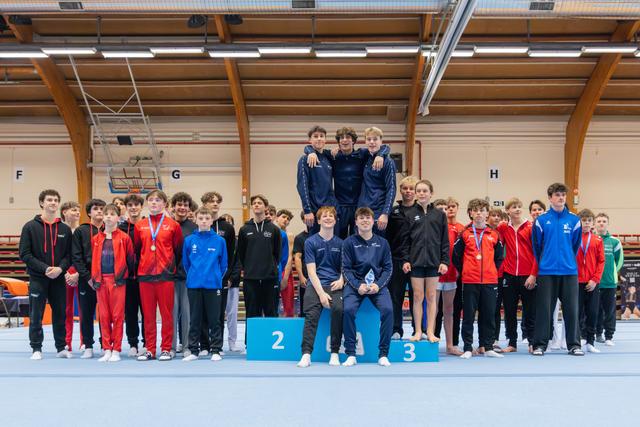 Young male gymnasts pose on podium, smiling proudly; teammates in red, black and blue surround them in a sports hall.