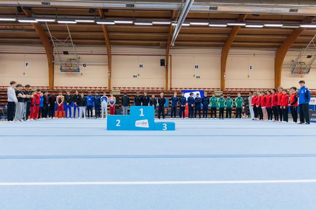 Teams line up around a 1-2-3 podium at an indoor gymnastics meet, awaiting a medal ceremony with calm anticipation.