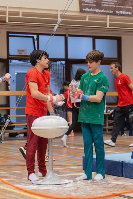 Two young gymnasts apply chalk before their event, the boy in green holding red rings while his teammate assists, both focused.