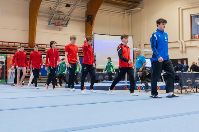 Young male gymnasts in red and blue jackets march across the floor exercise mat during a gymnastics competition.