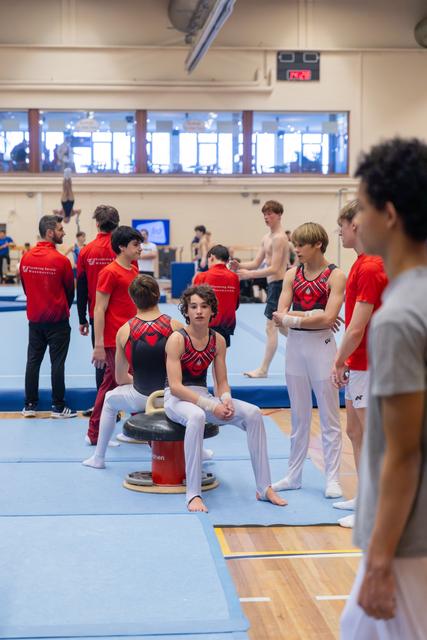 Young male gymnasts in red leotards chat and rest between routines on blue mats in a busy gymnasium.