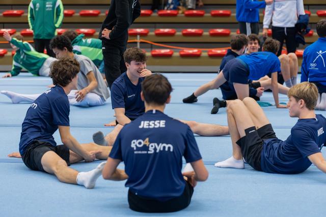 Young male gymnasts in navy GO4gym shirts sit on the floor mat laughing and chatting together between events.