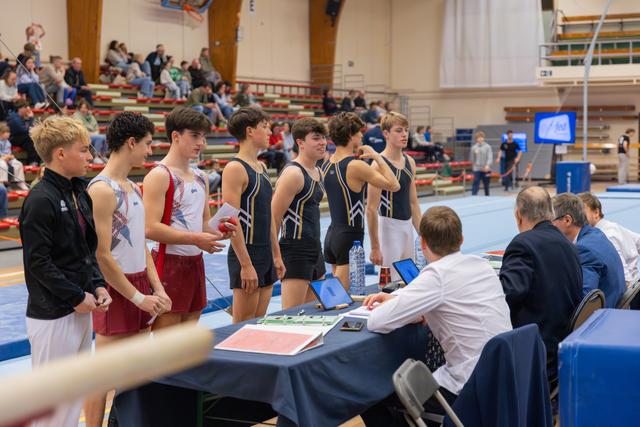 Young male gymnasts in leotards line up at the judges' table, looking focused and composed before their routine.