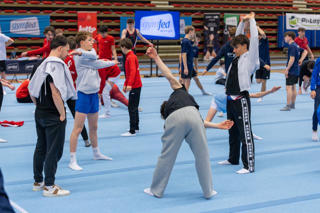 Young male gymnasts warm up on blue floor mats, stretching and reaching upward in a busy competition hall with Gymfed banners.