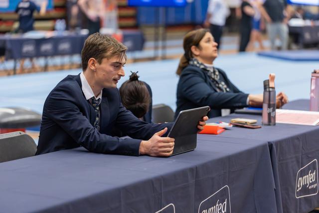 Young male judge in suit reviews tablet intently at judges' table during a Gymfed gymnastics competition.