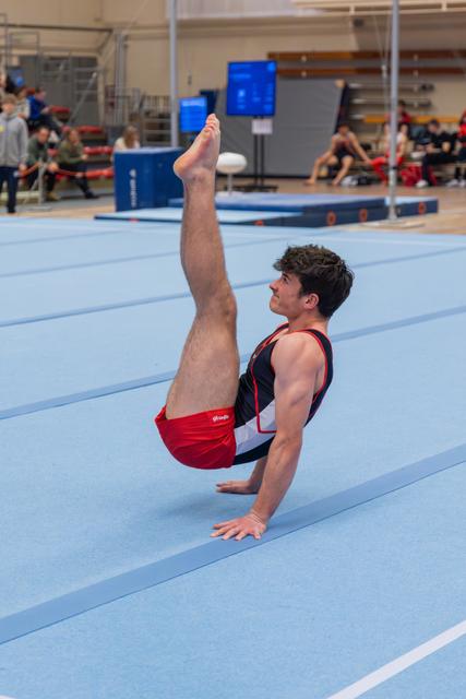 Male gymnast holds a V-sit position on the floor exercise mat, legs raised high, arms supporting, focused expression.