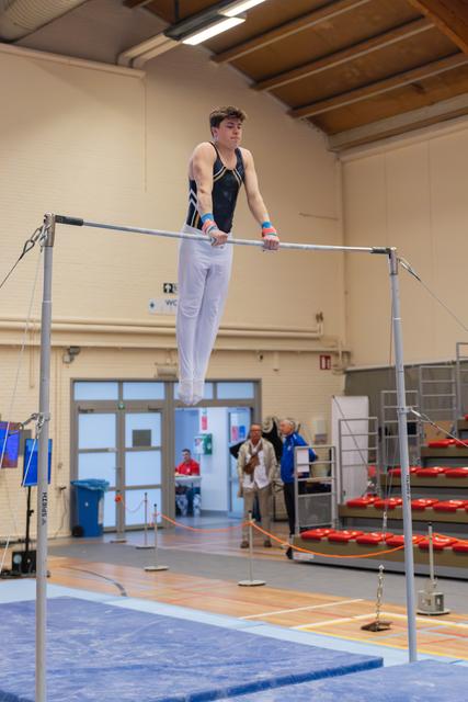 Young male gymnast grips the high bar with focused intensity, body suspended mid-air in a gymnastics hall.