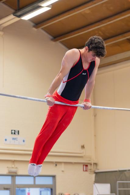 Young male gymnast grips the high bar, leaning forward with focused concentration in a gymnasium setting.