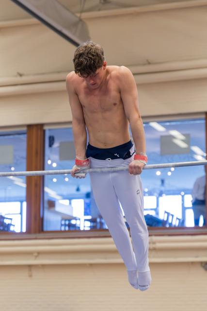 Young male gymnast grips parallel bars with focused intensity, shirtless in white trousers and red wrist guards, indoor gym.