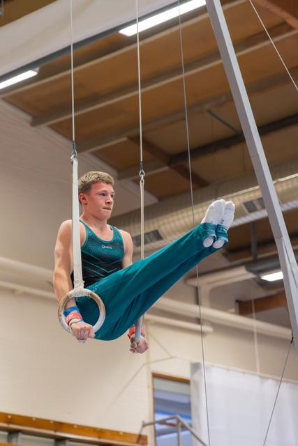 Young male gymnast performs a leg raise on still rings, focused expression, wearing teal leotard in an indoor gymnasium.