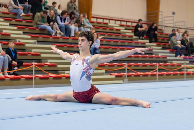 Young male gymnast performs a perfect split on the floor, arms extended, expression calm and focused, spectators in bleachers behind.