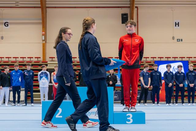 Young gymnast in red tracksuit stands on 3rd place podium as officials approach to present award in sports hall.