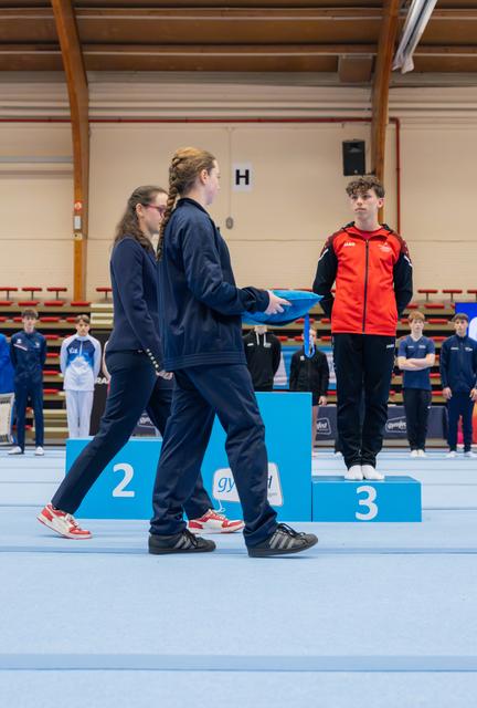 Two officials in navy tracksuits present a medal to a young gymnast in red standing on the third-place podium, expression serious.