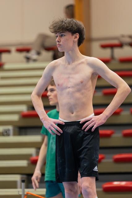 Shirtless teen gymnast stands with hands on hips, focused expression, in a gymnasium with red bleachers behind.