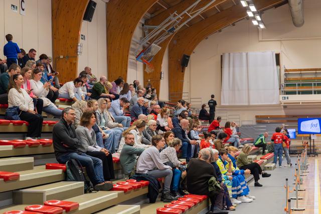 Crowd of spectators seated on red bleachers in an indoor sports hall, attentively watching an event on the court.