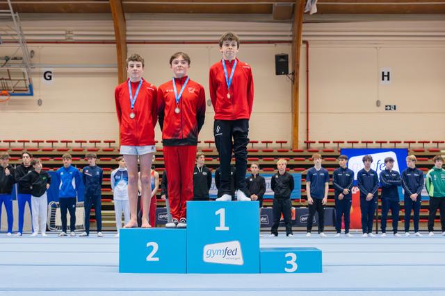 Three young male gymnasts stand proudly on a blue Gymfed podium, wearing red jackets and blue medals around their necks.