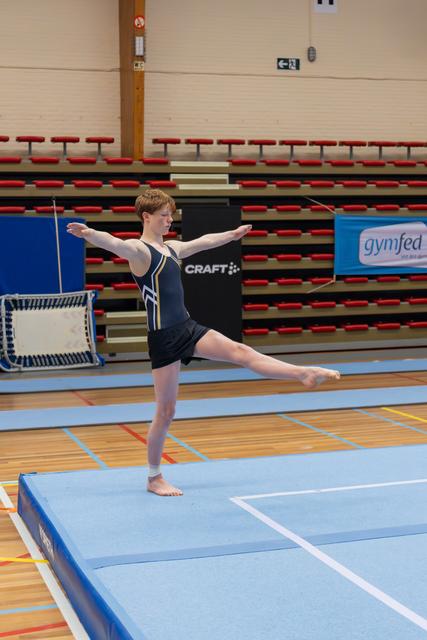 Young gymnast balances on one leg with arms extended, performing an arabesque on a blue floor mat in a sports hall.