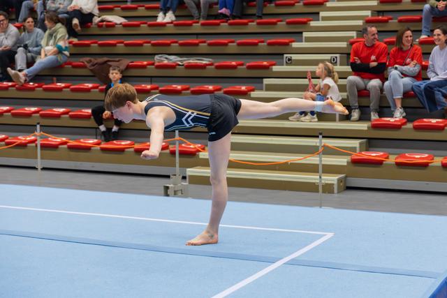 Young gymnast holds a precise arabesque balance on the floor mat, arms forward, leg raised, focused expression.
