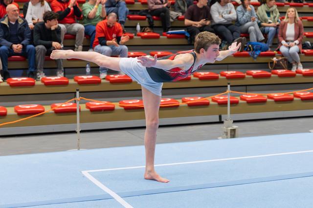 Teen male gymnast holds a precise arabesque on the floor, arms forward, focused expression, crowd watching in bleachers.