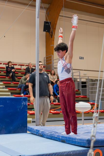 Young male gymnast stands tall on mat, arms raised holding rings, focused expression, coach watching in background.
