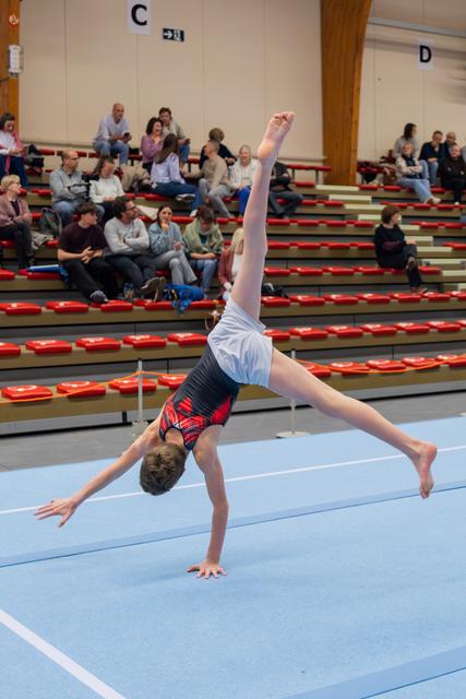 Young gymnast mid-cartwheel on blue floor mat, legs split wide, focused and controlled, audience watching in bleachers.