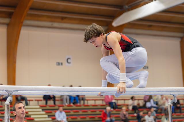 Young male gymnast crouches intently on parallel bars, concentration visible on his face, in a gymnasium with spectators.