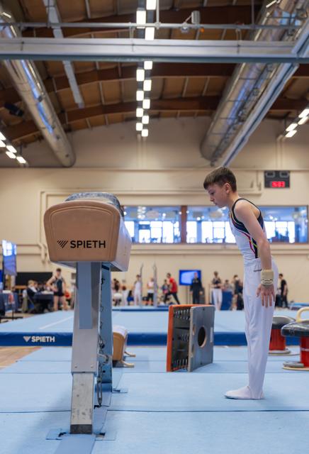 Young male gymnast stands focused, head bowed before a Spieth vault in a busy gymnastics hall.