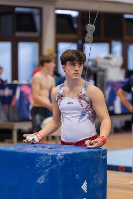 Young male gymnast stands focused at the vault, chalk on hands, wearing an Agiva leotard in a busy gym.