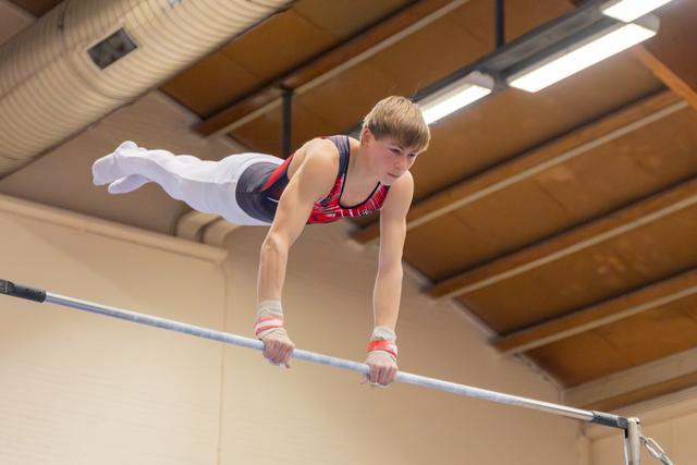 Determined young male gymnast grips the high bar mid-routine, body extended horizontally, in an indoor gymnasium.