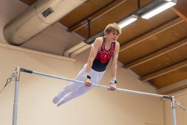 Focused young male gymnast performs on the horizontal bar, wearing wrist guards and a red-black leotard in an indoor gym.