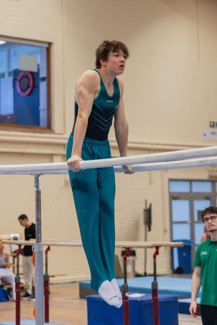 Focused young male gymnast balances on parallel bars, arms taut, looking ahead with concentration in an indoor gymnasium.