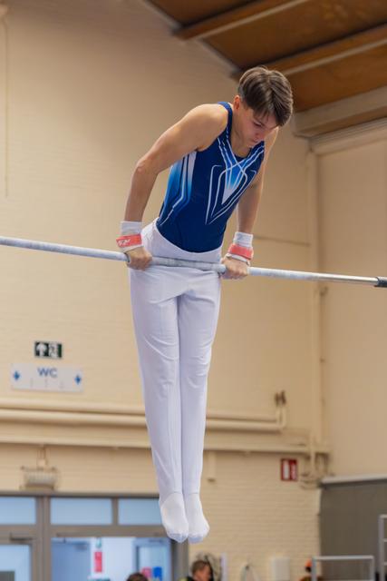 Young male gymnast in blue leotard holds a support position on parallel bars, head bowed in concentration.