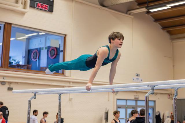 Young gymnast holds a focused planche position on parallel bars, body horizontal, determined expression in a gymnasium.