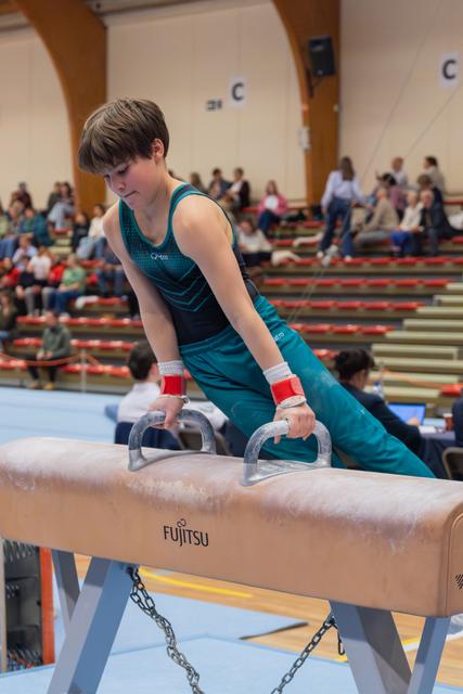 Focused young male gymnast grips pommel horse handles, leaning forward with intense concentration during a gymnastics competition.