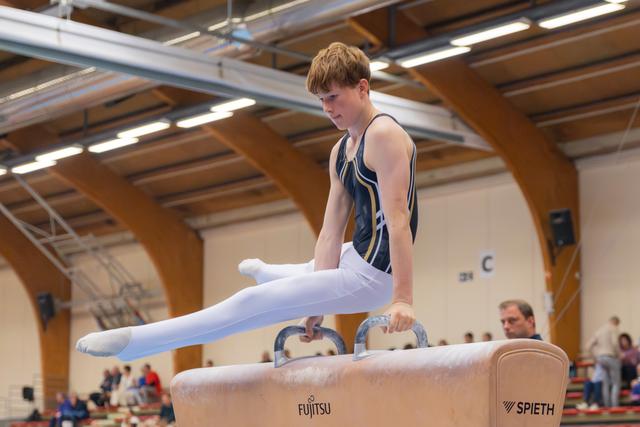 Focused teenage male gymnast performs on pommel horse, legs extended, intense concentration in a gymnasium.