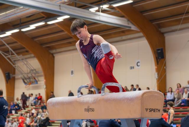 Focused young male gymnast performs on the pommel horse, leaning forward with intense concentration during a competition.