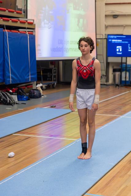Young gymnast stands composed on the floor mat, wearing a red leotard and wrist wraps, preparing for a routine.