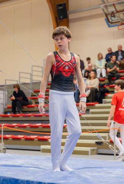 Young male gymnast in red leotard stands composed on blue mat, wrists taped, spectators watching in background.
