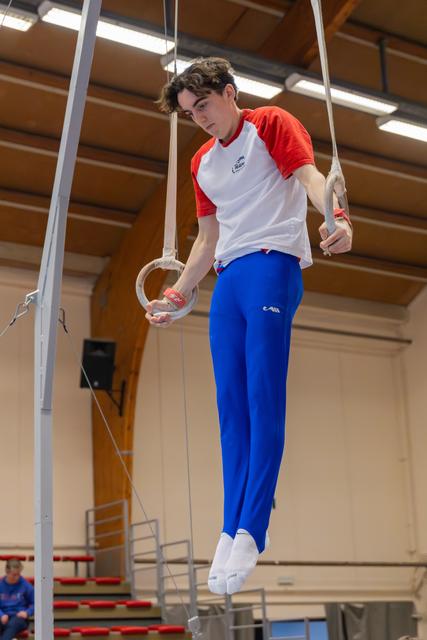 Focused teenage gymnast hangs on still rings in a gymnasium, wearing white and red shirt with blue trousers.