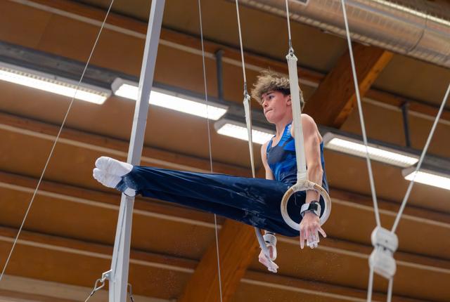Young male gymnast holds a tuck position on still rings, focused expression, competing in an indoor gymnasium.