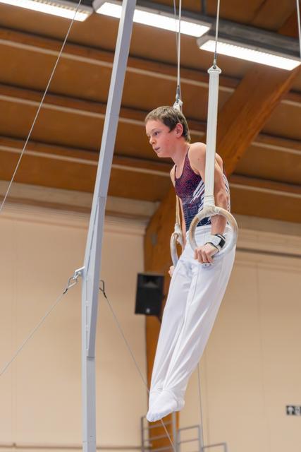 Young male gymnast holds still rings with intense focus, wearing white pants and red leotard in an indoor gymnasium.