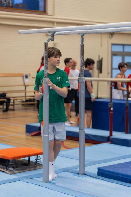 Young male gymnast in green shirt grips parallel bars, looking intently sideways in a busy gymnastics hall.