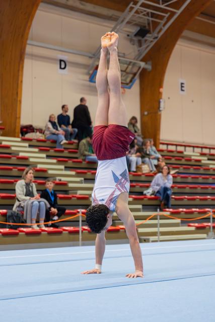 Young gymnast performs a handstand on the floor exercise mat, legs perfectly straight, in a sports arena with spectators.