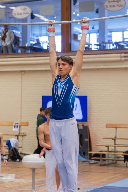 Young male gymnast hangs from the high bar, focused expression, wearing blue leotard and white pants in an indoor gym.