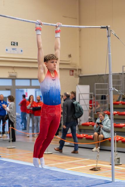 Focused teenage male gymnast hangs on the high bar with intense concentration, wearing a red and blue leotard indoors.