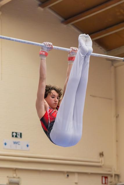 Young male gymnast hangs from high bar in a tight pike position, focused expression, wearing red leotard and white tights.