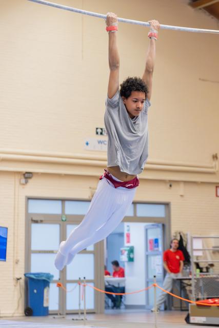 Young gymnast hangs from a high bar with intense focus, wearing white leggings and red wristbands in an indoor gym.
