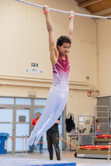 Young male gymnast hangs from the high bar with focused concentration, wearing a maroon and white leotard indoors.